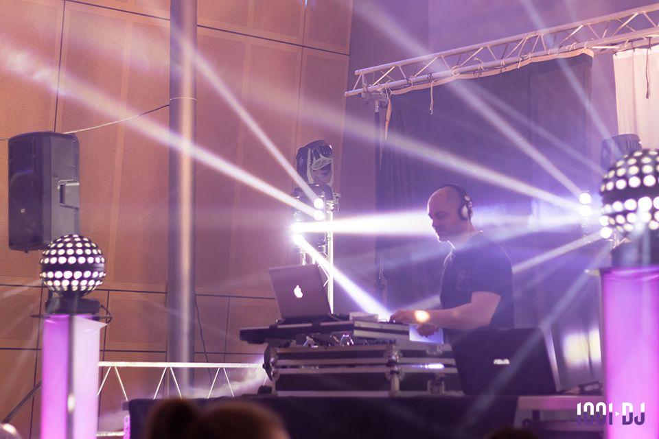 Un DJ mixant sur une scène éclairée par des projecteurs et des faisceaux lumineux dans une salle.