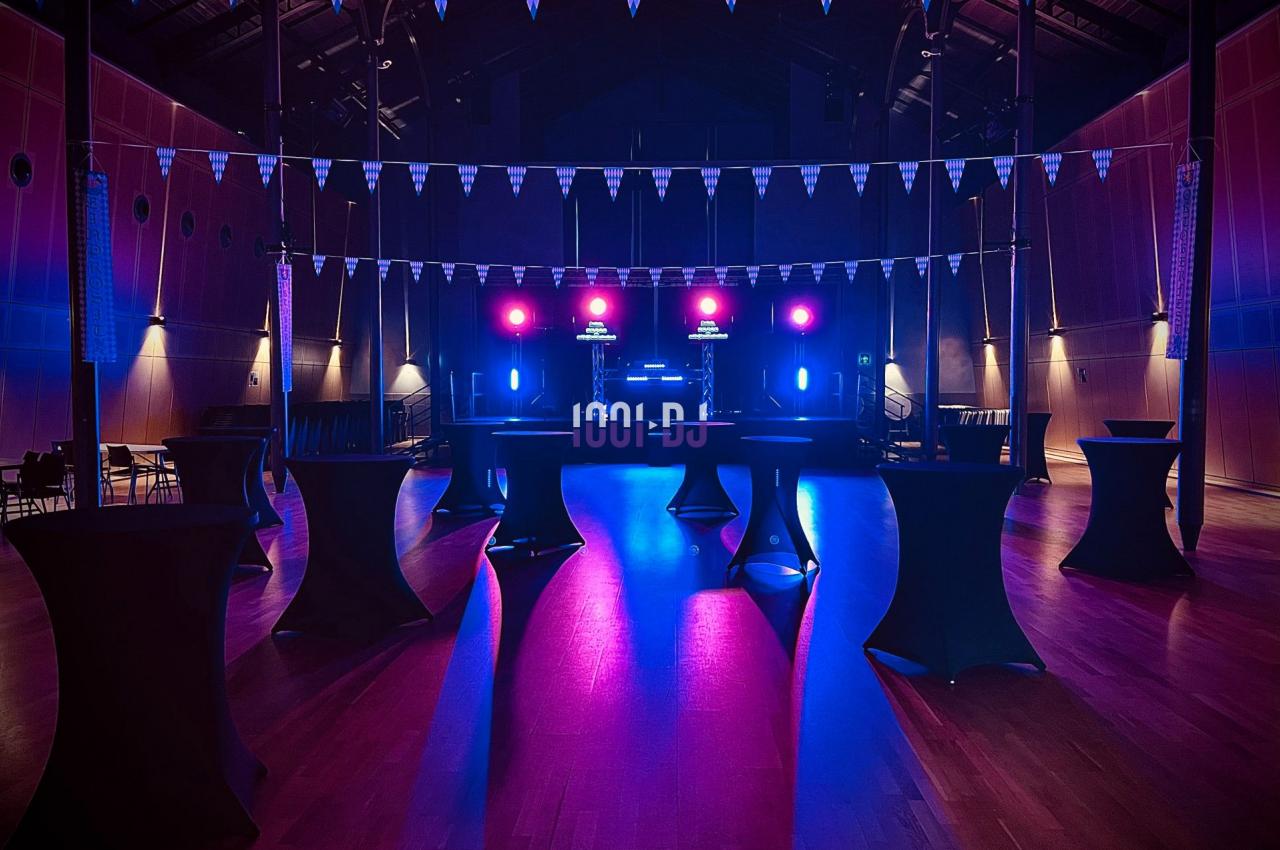 Salle de réception avec éclairage coloré, tables hautes noires et scène équipée de lumières et matériel audio.