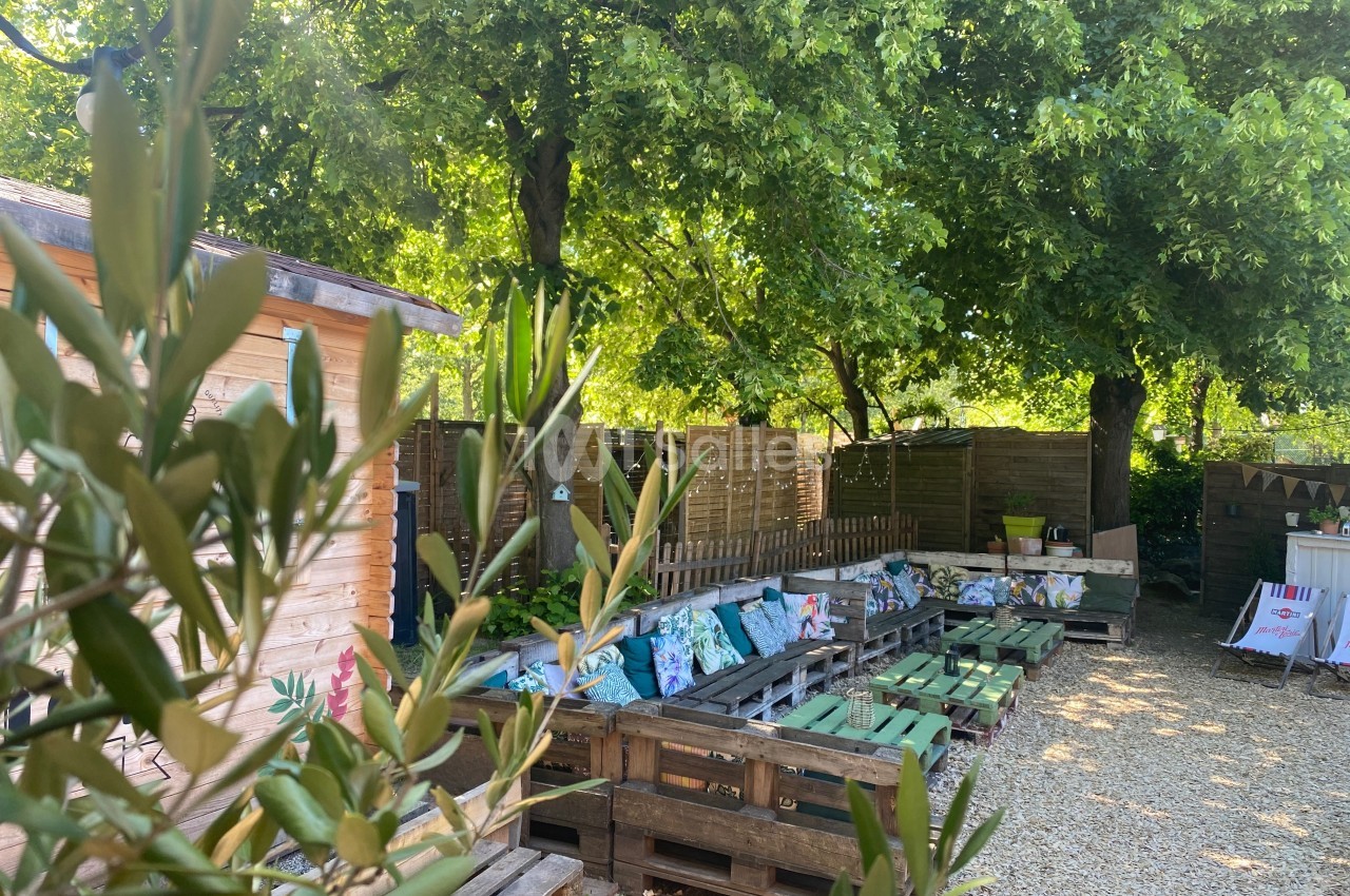 Espace extérieur ombragé avec bancs en palettes, coussins colorés, cabanon en bois et arbres verdoyants.