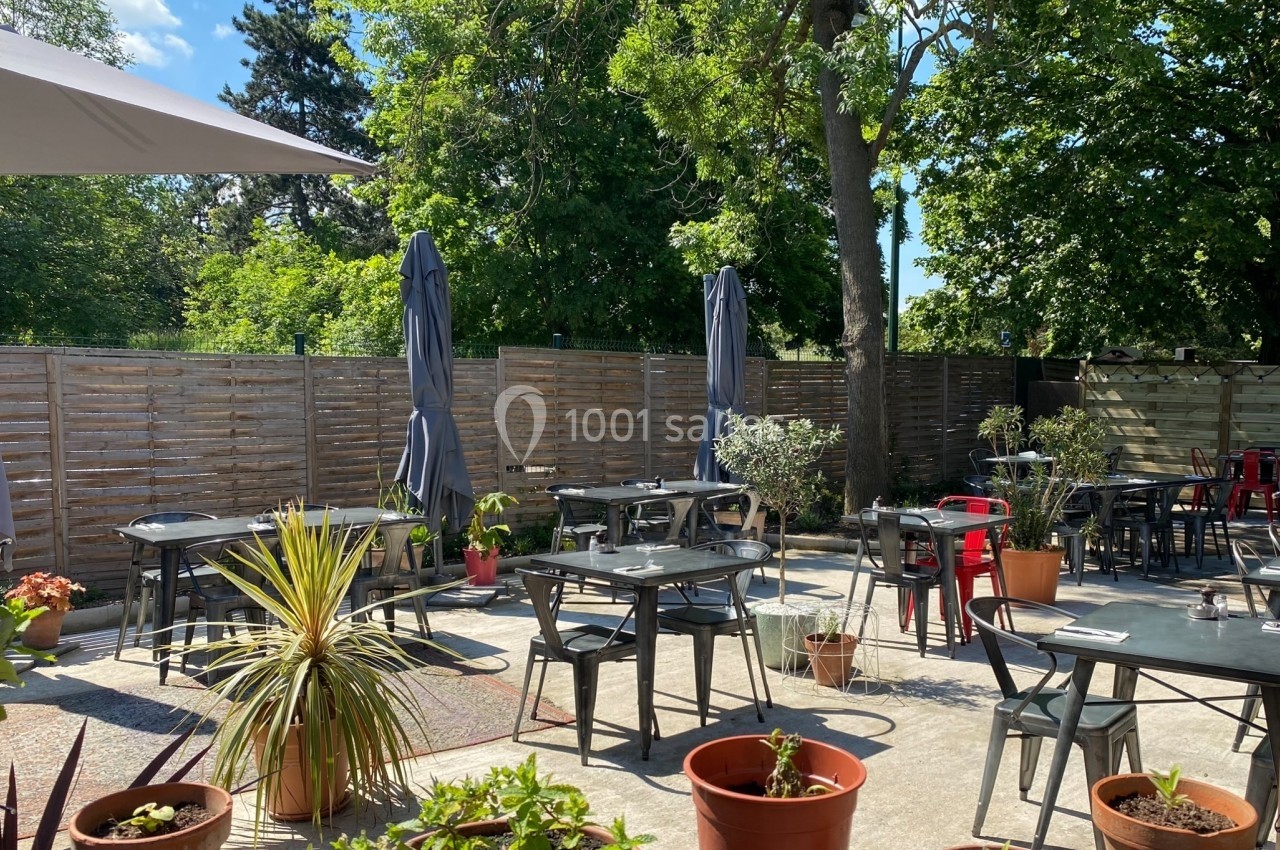 Terrasse extérieure avec tables, chaises, parasols et plantes en pots, entourée de verdure et d'une clôture en bois.
