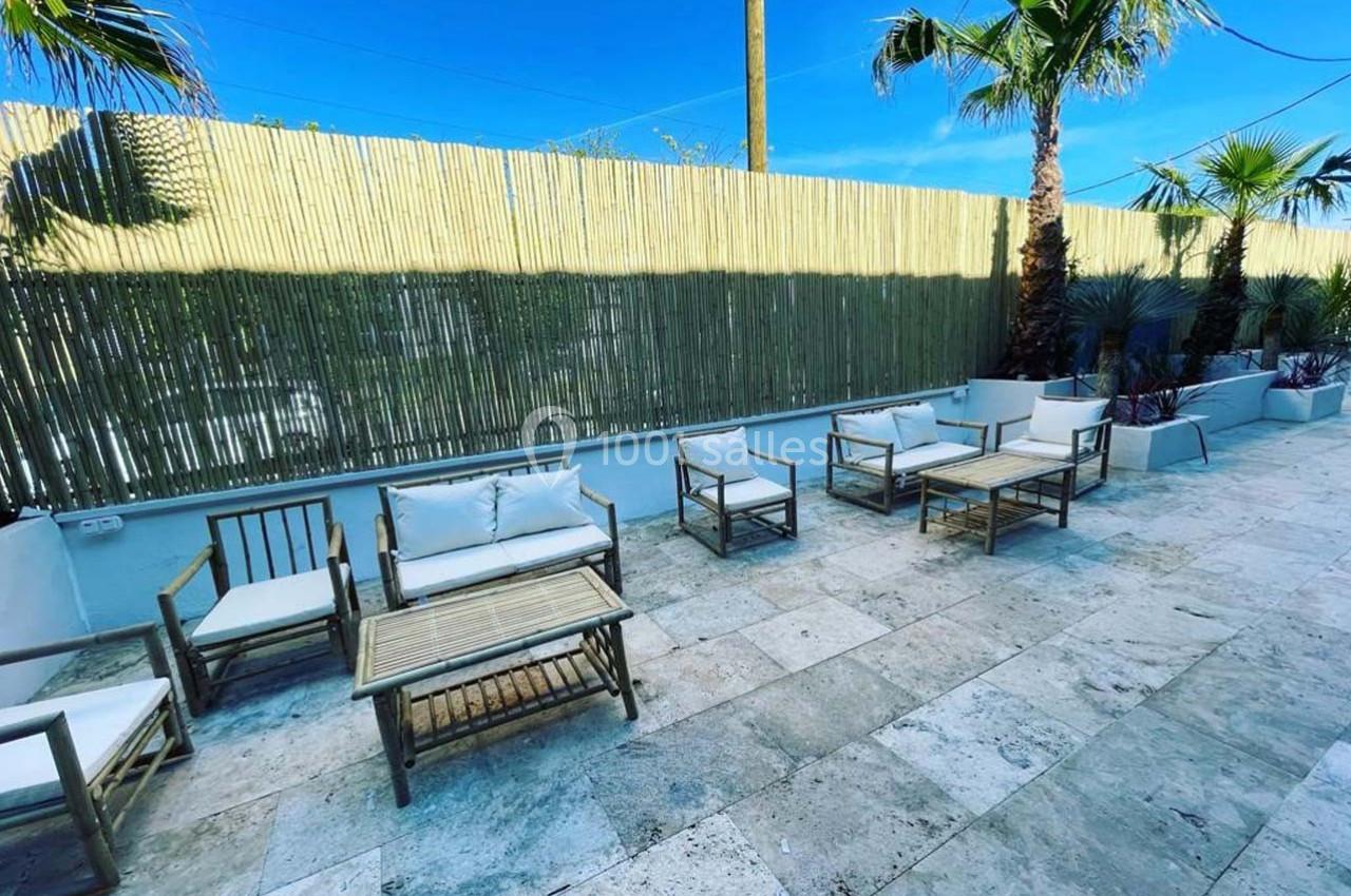 Mobilier de jardin en bois avec coussins blancs disposé sur une terrasse en pierre, entourée de palmiers et d'une clôture en…