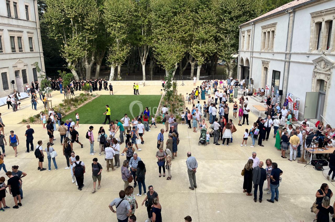 Foule rassemblée dans une cour extérieure bordée d'arbres et de bâtiments historiques par une journée ensoleillée.