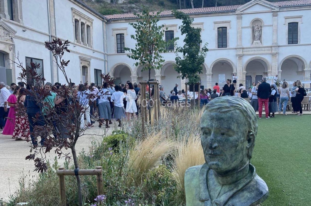 Cour intérieure avec des plantes, une statue en premier plan et un groupe de personnes rassemblées près d'un bâtiment ancien.