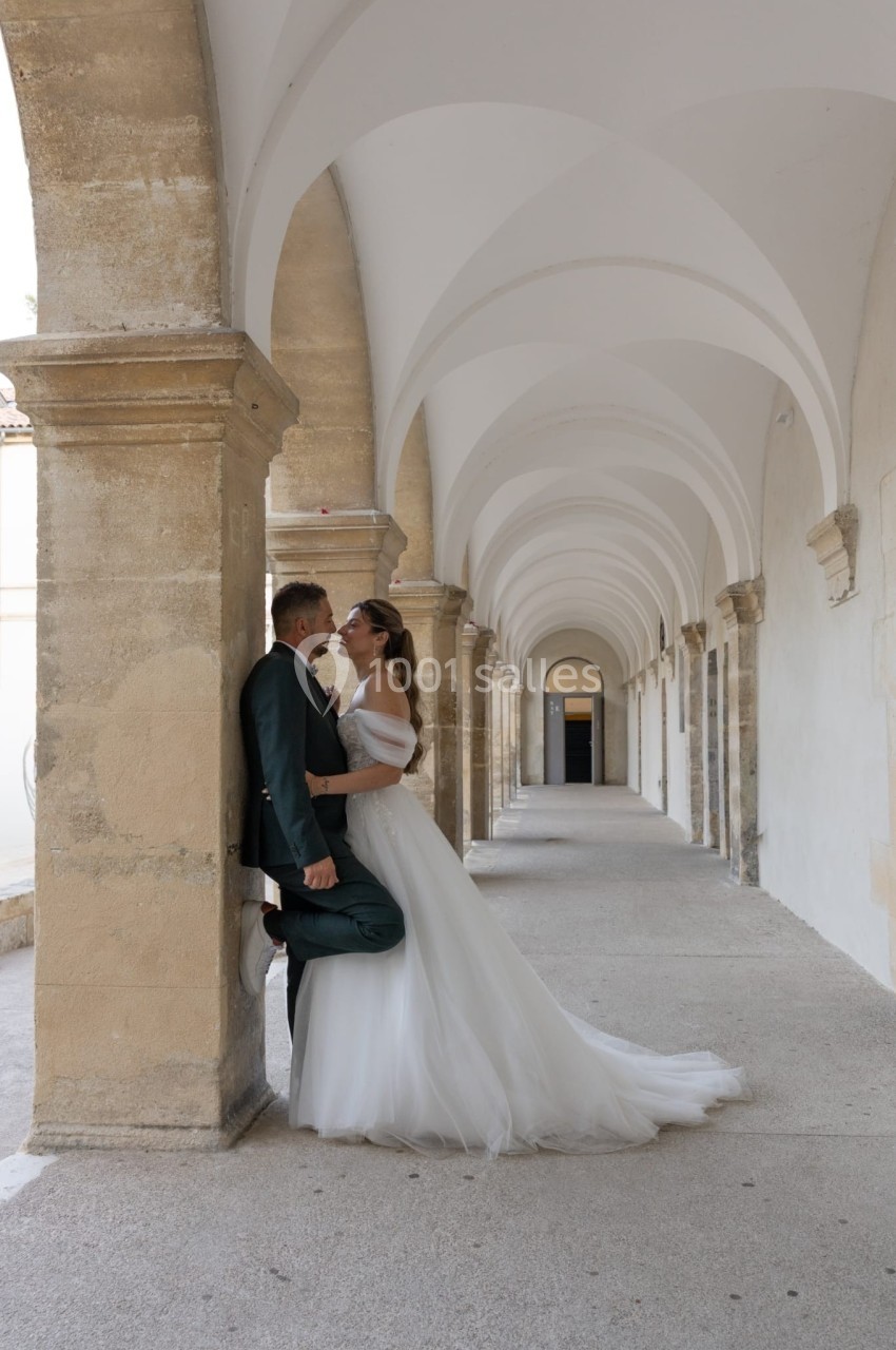 Un couple en tenue de mariage pose sous une galerie voûtée en pierre, dans un cadre architectural élégant.