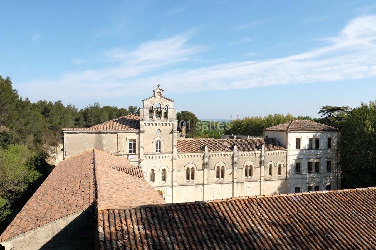 Vue d'un bâtiment historique en pierre avec toits en tuiles, entouré de végétation et sous un ciel dégagé.