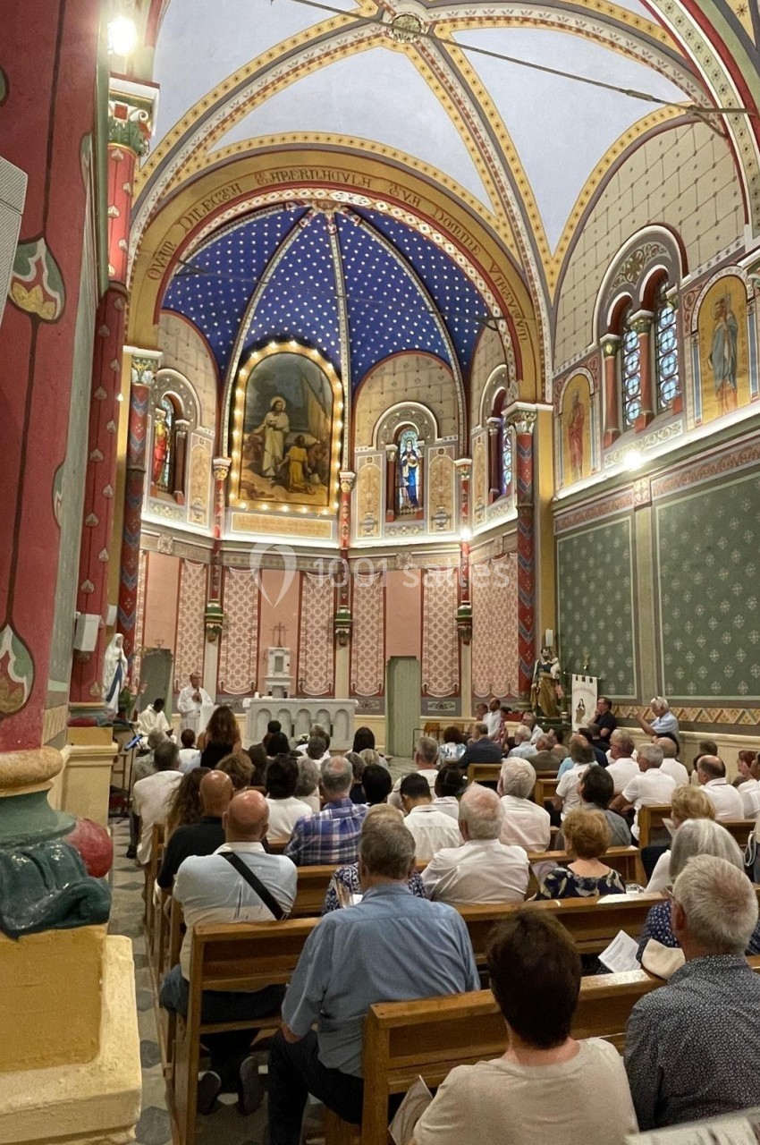 Intérieur d'une église avec des fidèles assis, des fresques colorées et un prêtre officiant près de l'autel.