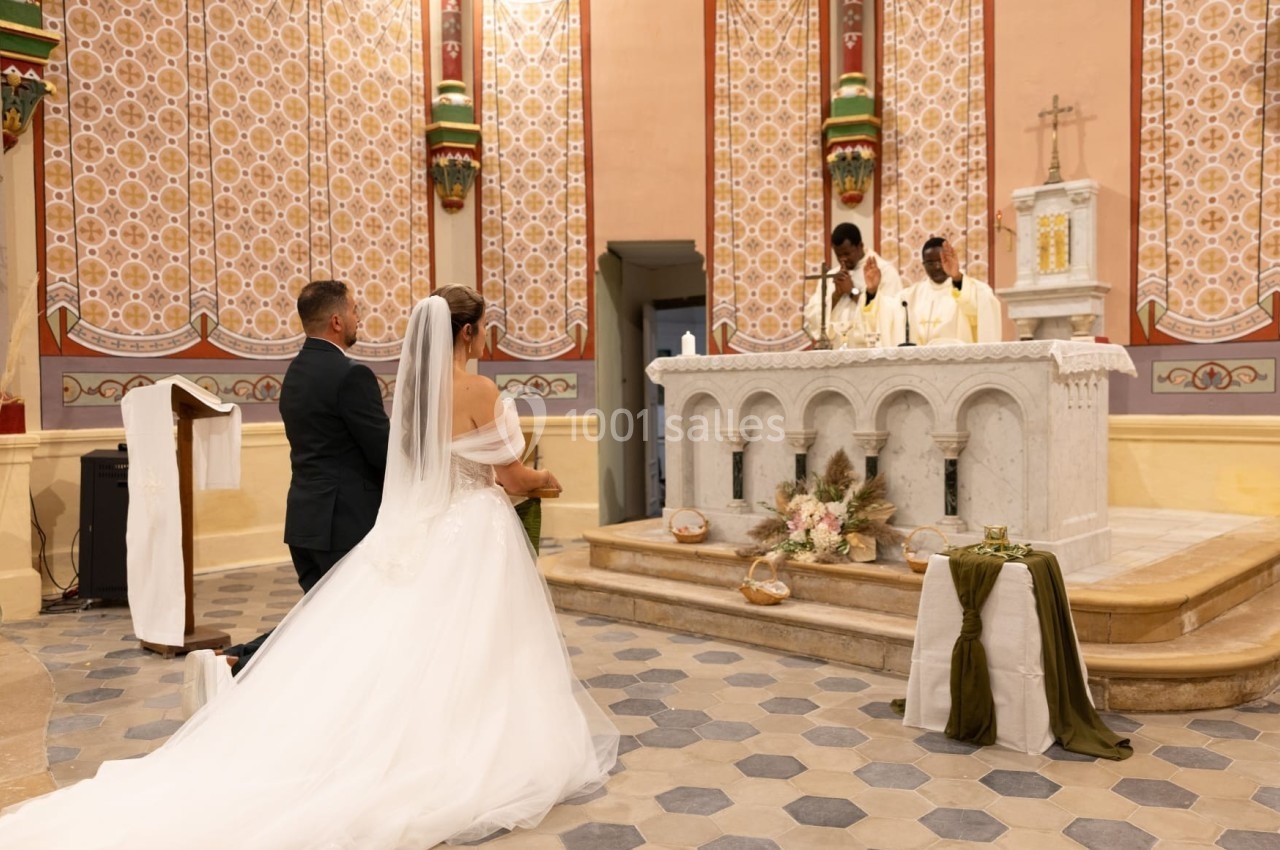 Un couple en tenue de mariage s'agenouille devant un autel dans une église, avec deux prêtres officiant.