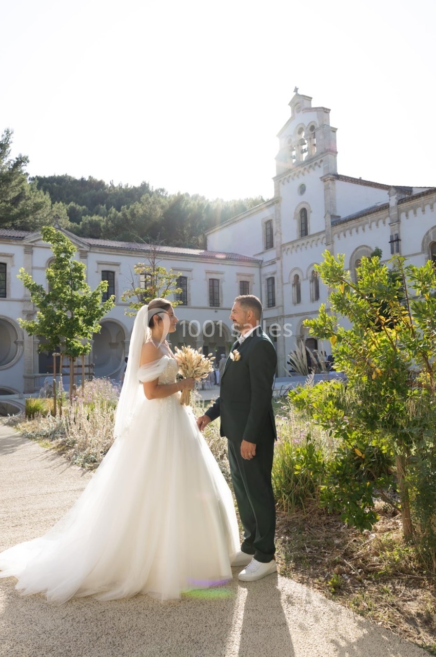 Un couple en tenue de mariage se tient face à face dans un jardin devant un bâtiment ancien sous un ciel ensoleillé.