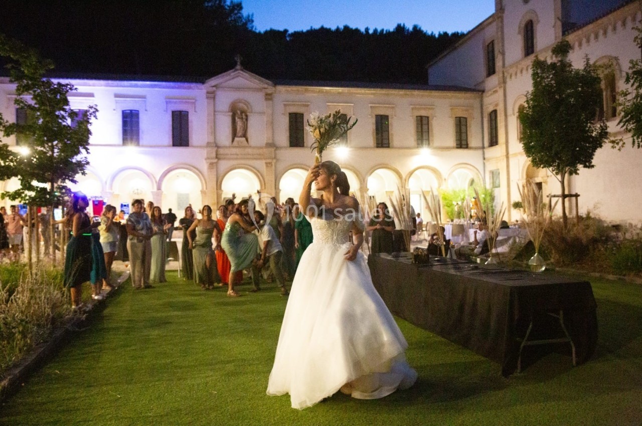Une mariée lance son bouquet devant des invités rassemblés dans le jardin d'un bâtiment éclairé en soirée.