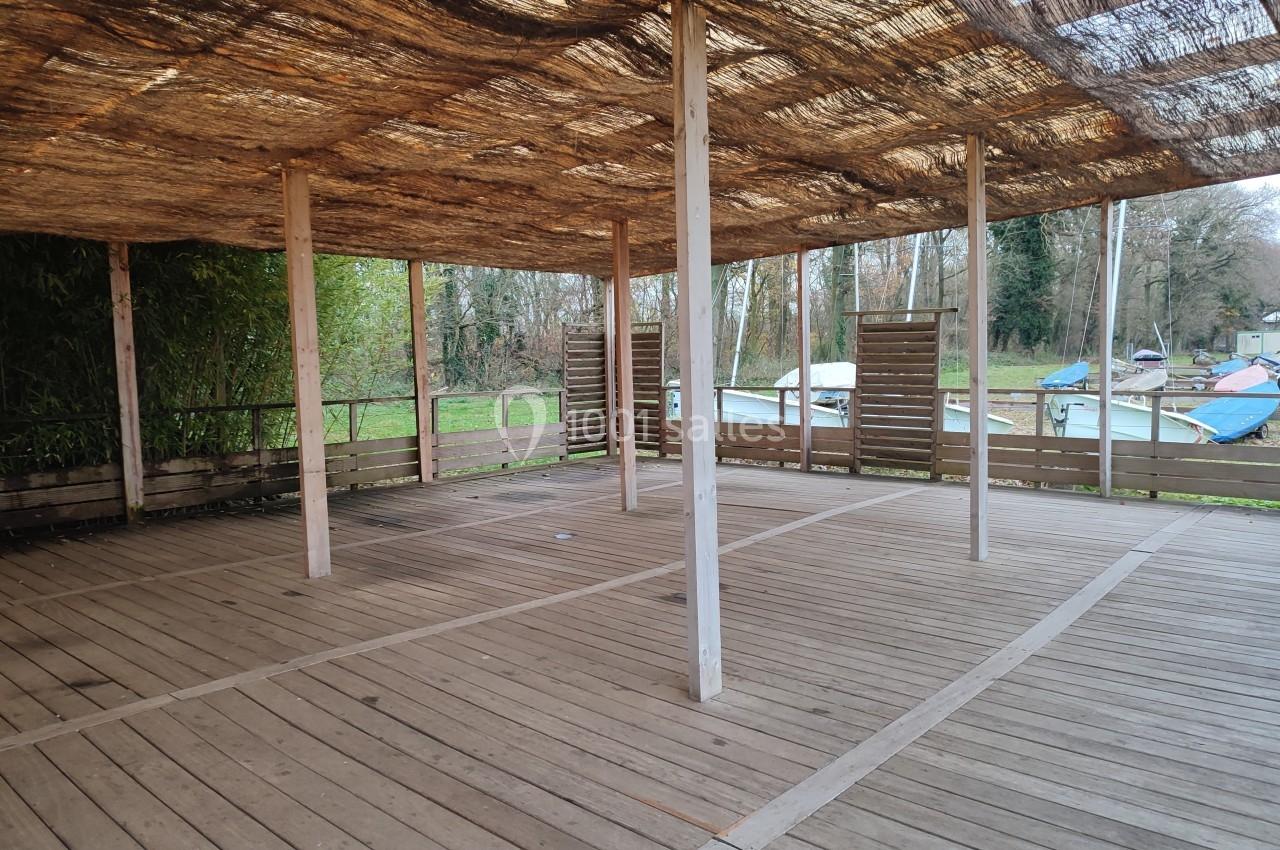 Structure en bois ouverte avec un toit en canisse, entourée de végétation et située près de bateaux amarrés.