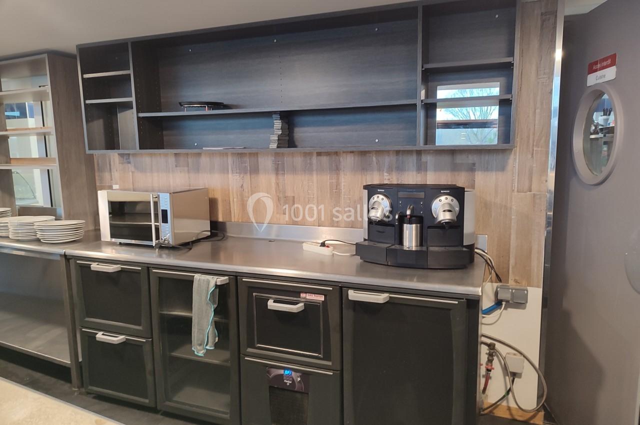 Cuisine équipée avec étagères vides, micro-ondes, machine à café et rangements noirs sous un plan de travail en inox.