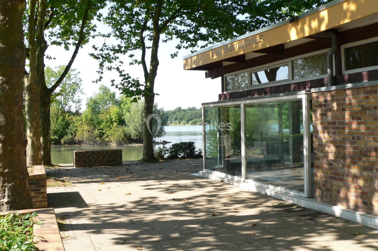 Bâtiment en briques avec grandes baies vitrées, entouré d'arbres, près d'un plan d'eau sous un ciel dégagé.