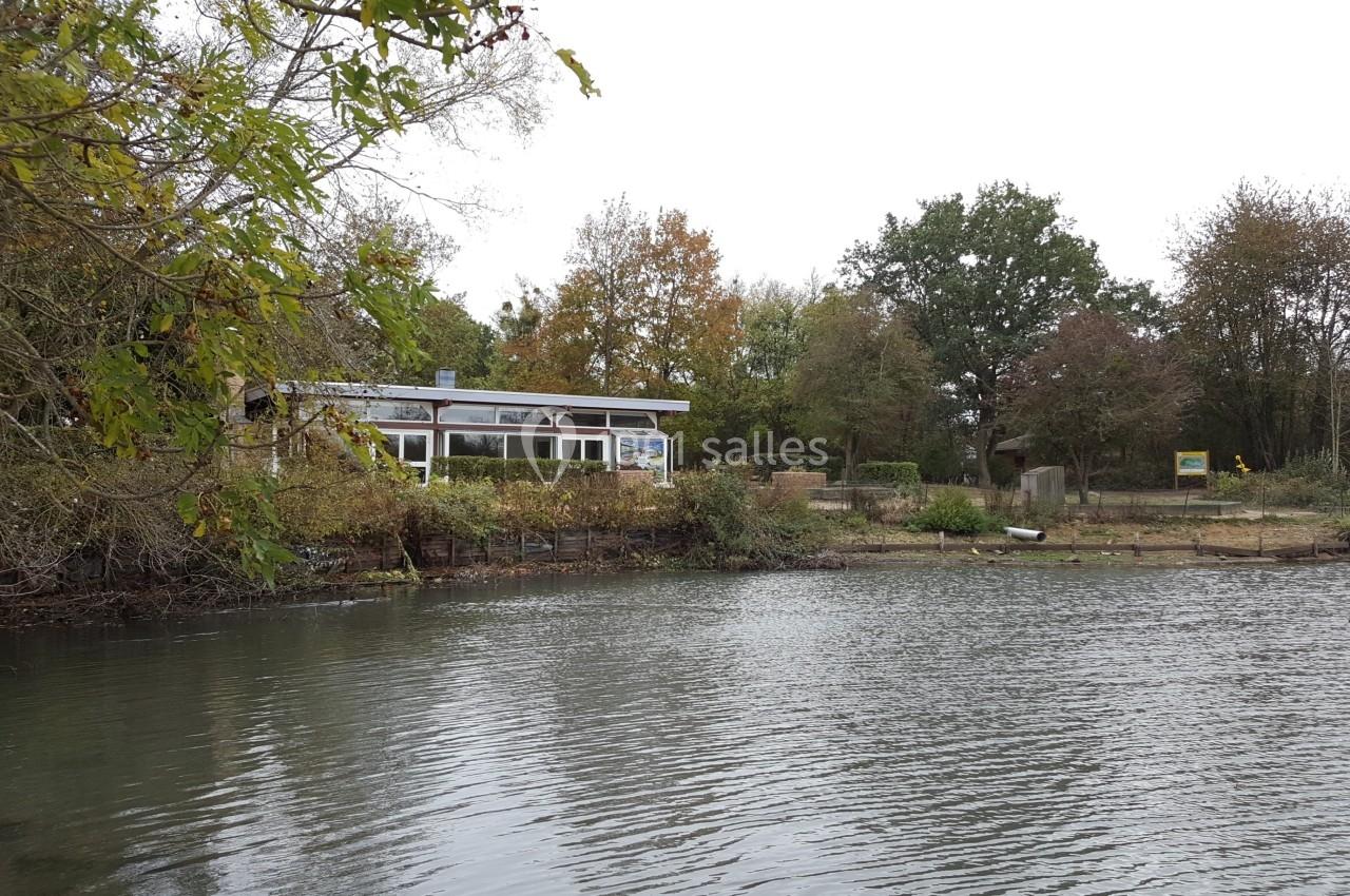 Vue d'un bâtiment au bord d'un étang entouré d'arbres en automne, avec une rive légèrement dégagée.