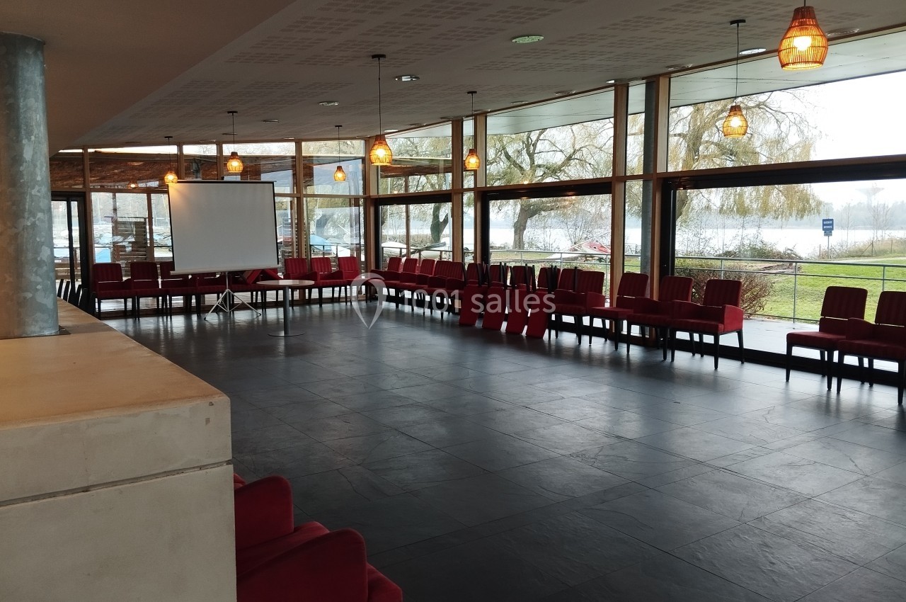 Salle lumineuse avec grandes baies vitrées, chaises rouges alignées et écran de projection au fond.