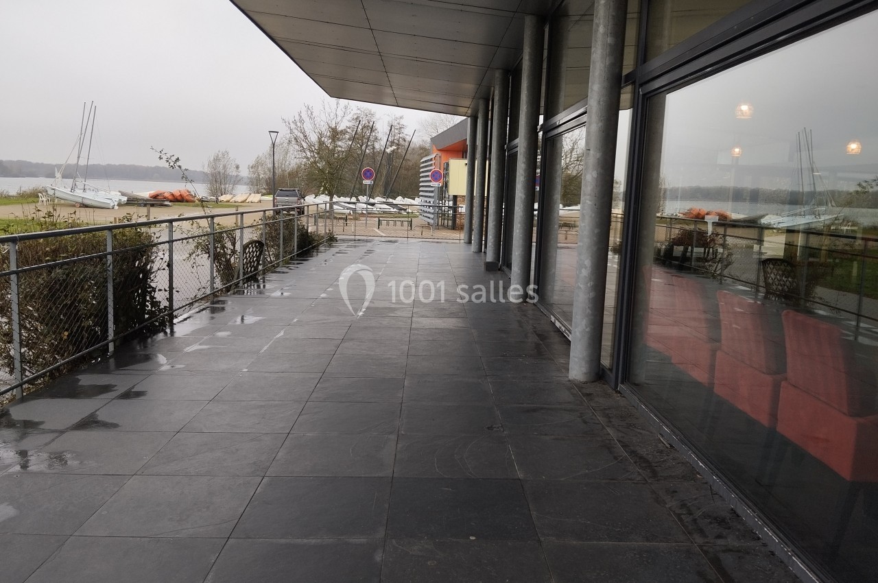 Terrasse couverte avec sol en dalles sombres, vue sur un lac, des voiliers et des arbres par temps nuageux.