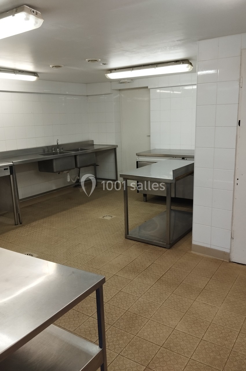Cuisine professionnelle vide avec plans de travail en inox, murs carrelés blancs et sol en carrelage beige.