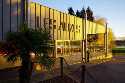 Bâtiment en briques avec de grandes baies vitrées, entouré d'arbres, près d'un plan d'eau sous un ciel dégagé.