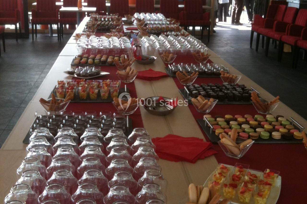 Table dressée avec des verrines, macarons, biscuits et verres, dans une salle lumineuse avec des invités en arrière-plan.