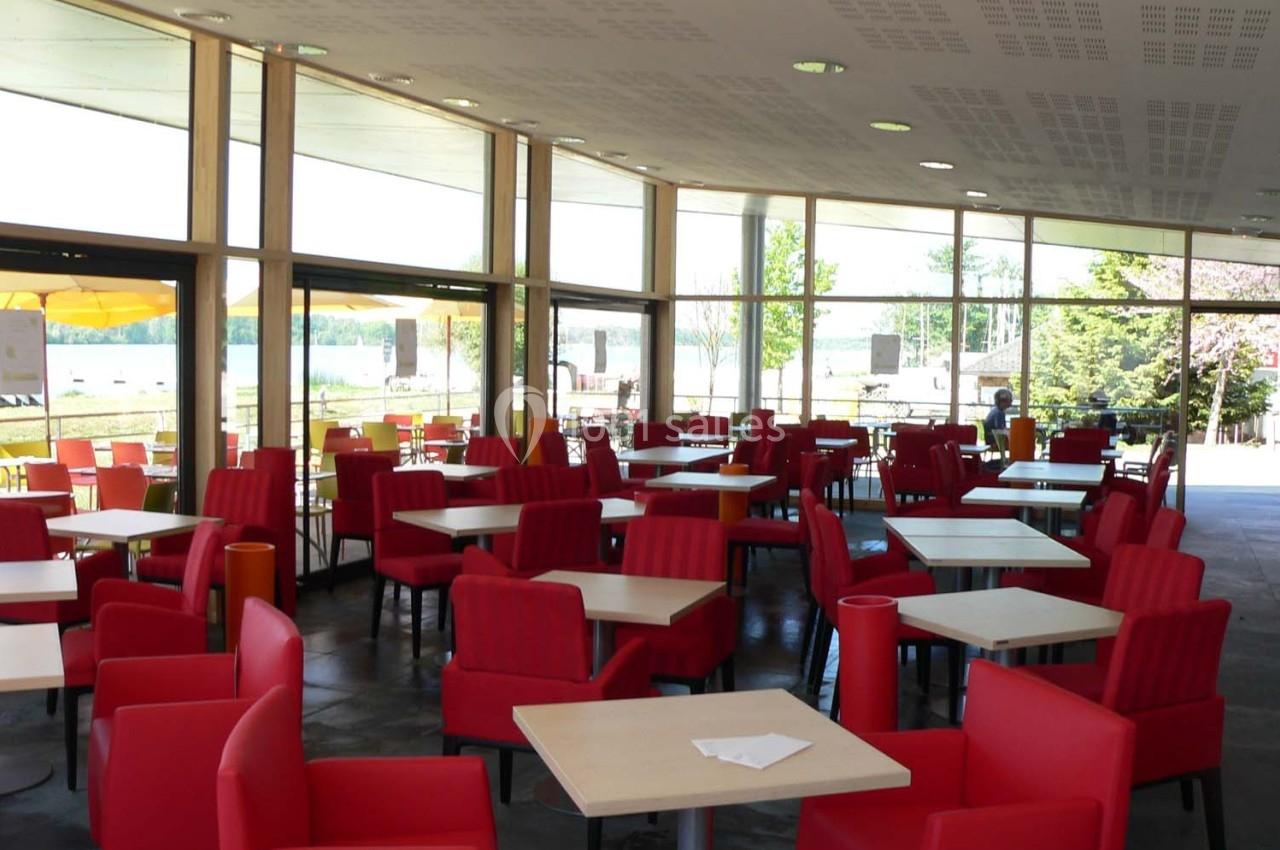 Salle lumineuse avec de grandes baies vitrées, tables carrées et chaises rouges disposées de manière espacée.