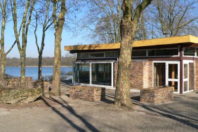 Bâtiment en briques avec de grandes baies vitrées, entouré d'arbres, près d'un plan d'eau sous un ciel dégagé.