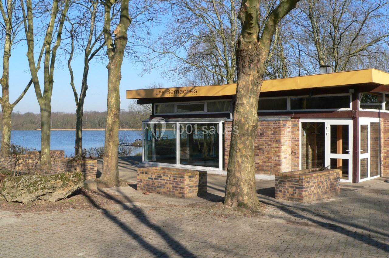 Bâtiment moderne en briques avec de grandes baies vitrées, entouré d'arbres près d'un lac sous un ciel dégagé.