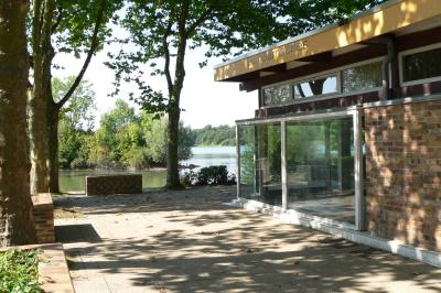 Bâtiment en briques avec de grandes baies vitrées, entouré d'arbres, près d'un plan d'eau sous un ciel dégagé.