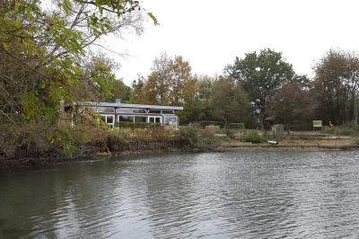 Bâtiment en briques avec de grandes baies vitrées, entouré d'arbres, près d'un plan d'eau sous un ciel dégagé.