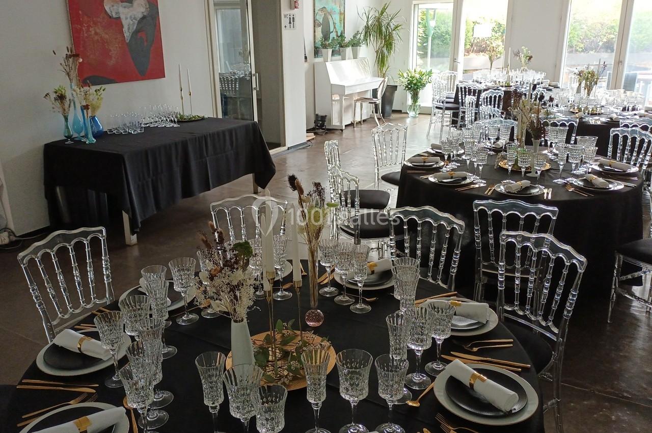Salle de réception avec tables rondes dressées, nappes noires, chaises transparentes et décoration florale.