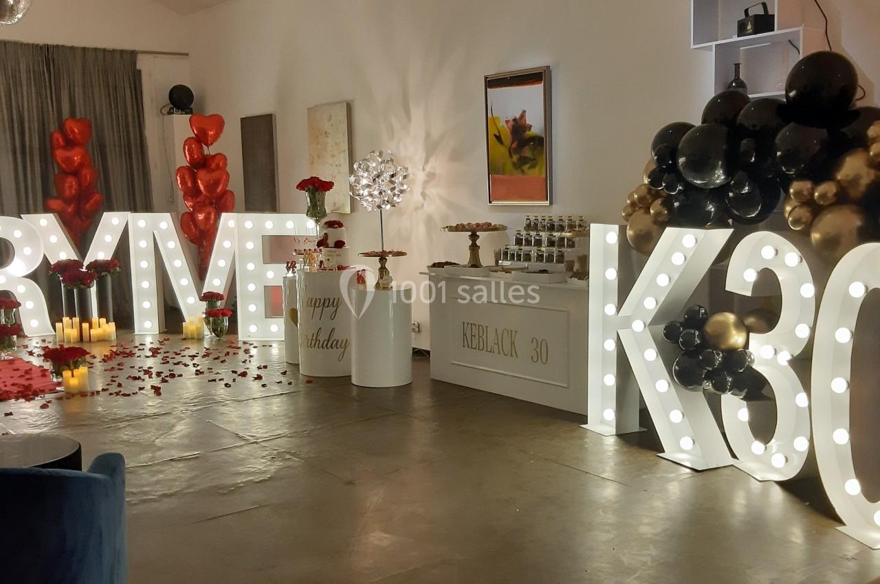 Salle décorée pour une fête avec ballons noirs et dorés, lettres lumineuses, bougies et pétales de rose au sol.