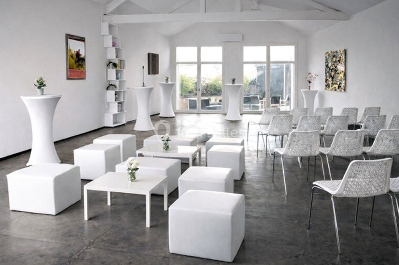 Salle lumineuse aménagée avec des chaises, des tables basses et des tabourets blancs, décorée de plantes et d'œuvres murales.