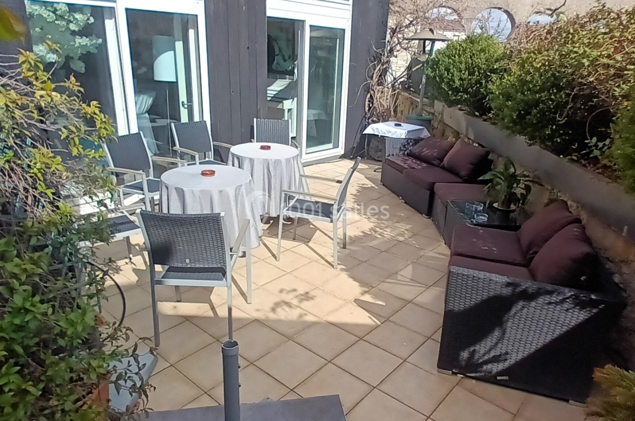 Terrasse aménagée avec tables, chaises et canapés entourée de plantes, située devant une baie vitrée.