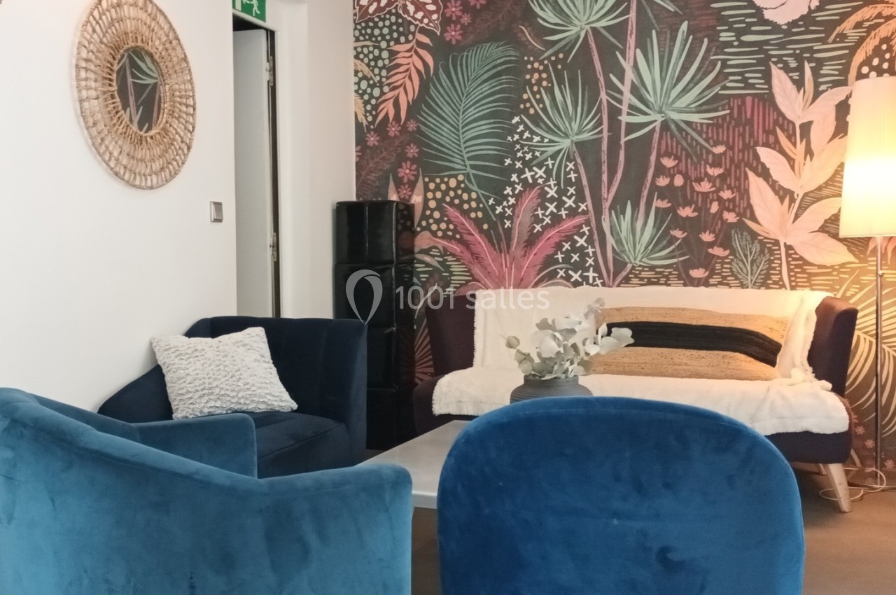 Salon cosy avec fauteuils en velours bleu, canapé décoré de coussins, table basse et mur orné d’un motif végétal coloré.