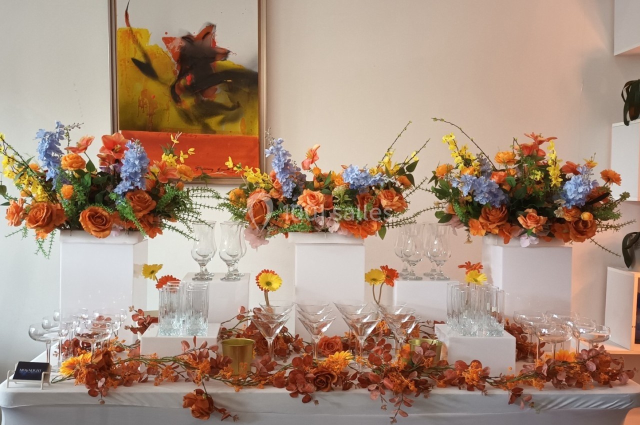 Table décorée avec des fleurs orange, jaunes et bleues, entourant des verres en cristal, dans un cadre élégant.