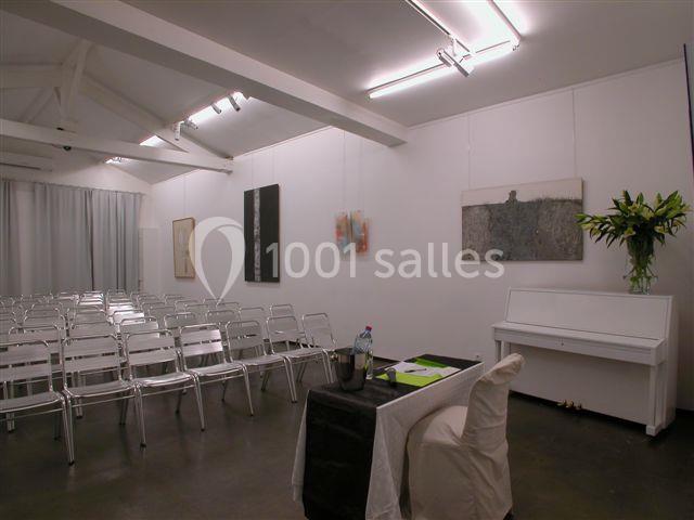 Salle lumineuse avec des chaises alignées, un piano blanc, des tableaux accrochés et une table préparée pour une…