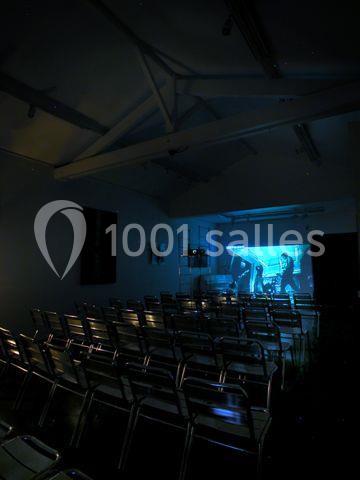 Salle sombre avec des rangées de chaises métalliques vides face à un écran projetant une image lumineuse.