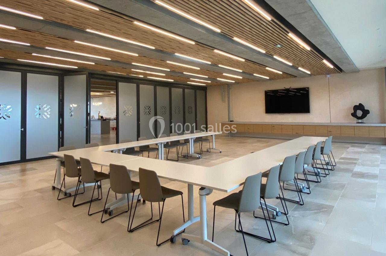 Salle de réunion moderne avec tables en U, chaises beige, plafond en bois et écran mural.