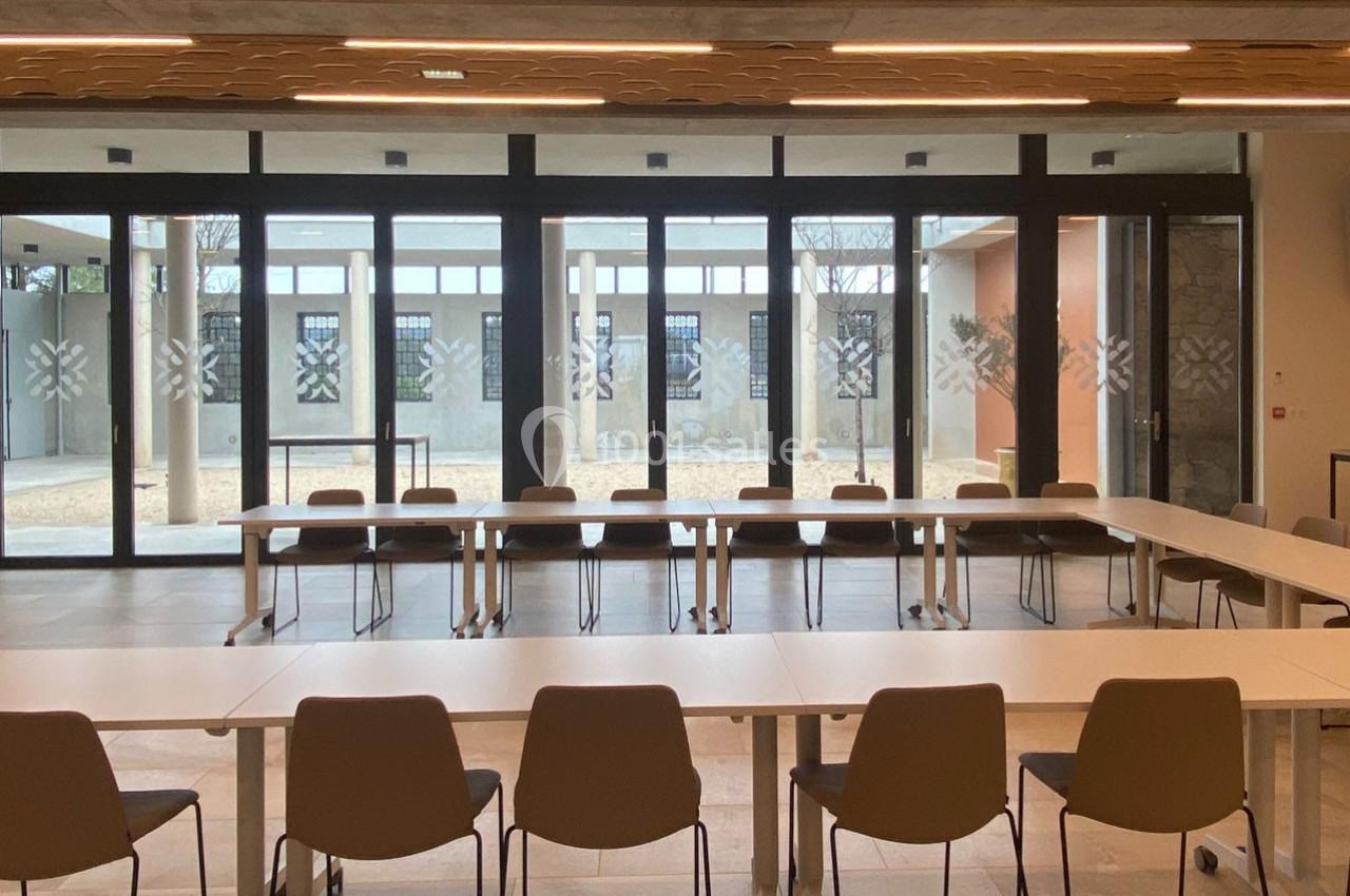 Salle de réunion lumineuse avec des tables disposées en U, des chaises et une grande baie vitrée donnant sur une cour.