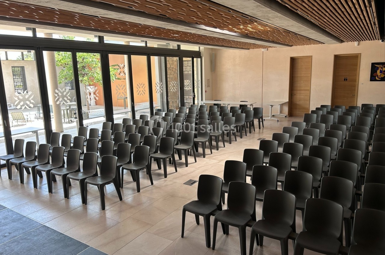 Salle lumineuse avec rangées de chaises noires alignées face à une estrade, grandes baies vitrées en arrière-plan.