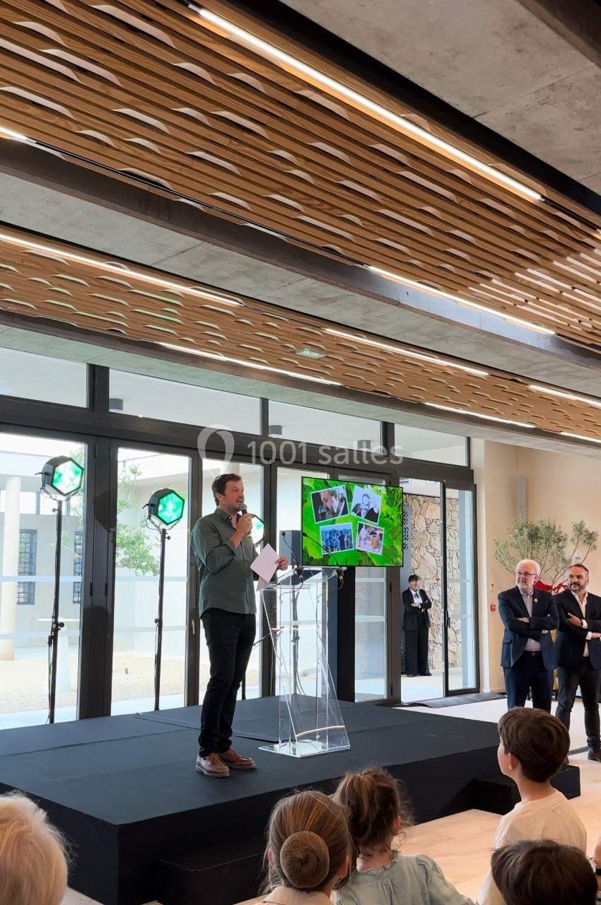 Un homme parle sur une scène avec un micro, devant un public, dans une salle moderne avec de grandes baies vitrées.