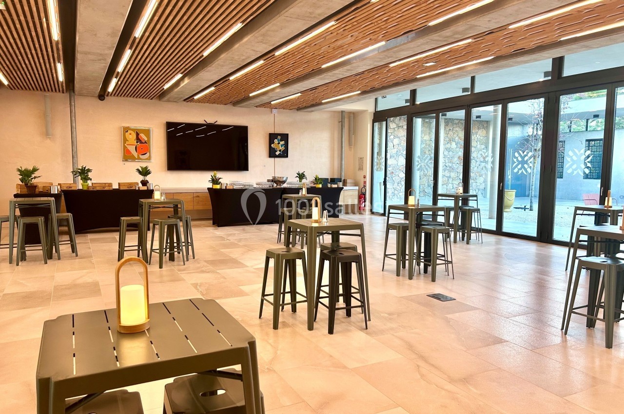 Salle lumineuse avec tables hautes, tabourets, éclairage moderne et grandes baies vitrées donnant sur une terrasse.