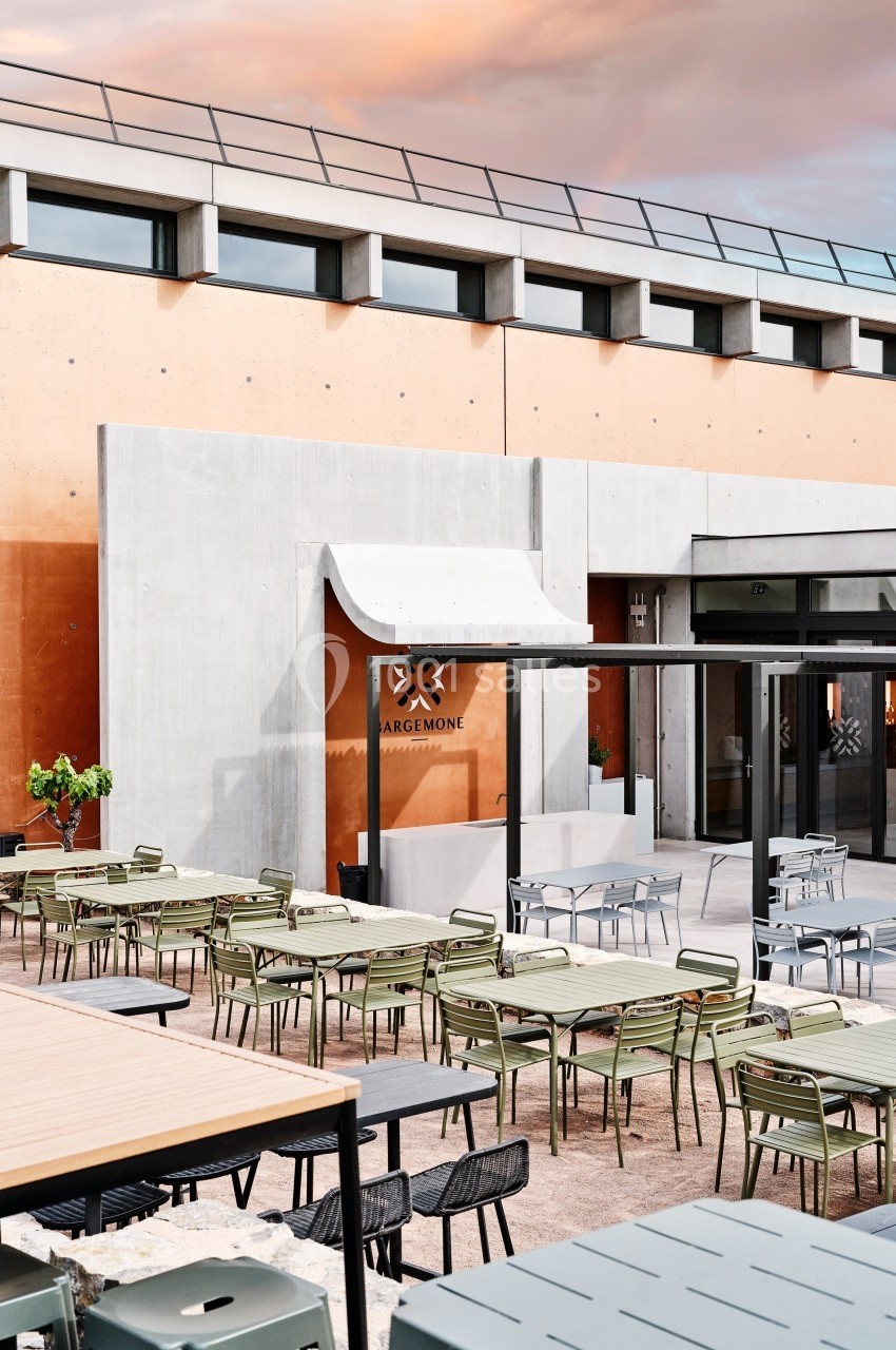 Terrasse extérieure avec tables et chaises modernes devant un bâtiment en béton et façade orange sous un ciel coloré.