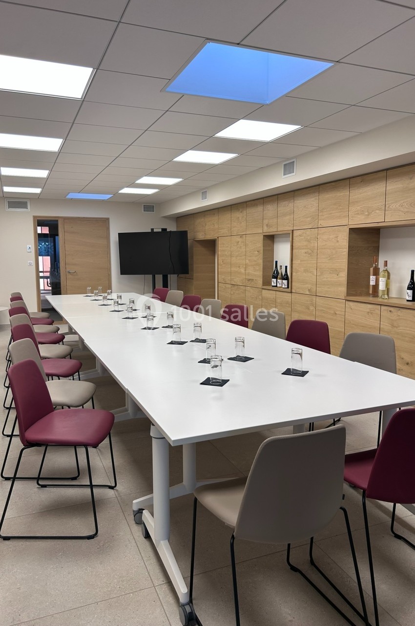 Salle de réunion avec une grande table blanche entourée de chaises, des verres disposés et un écran au fond.