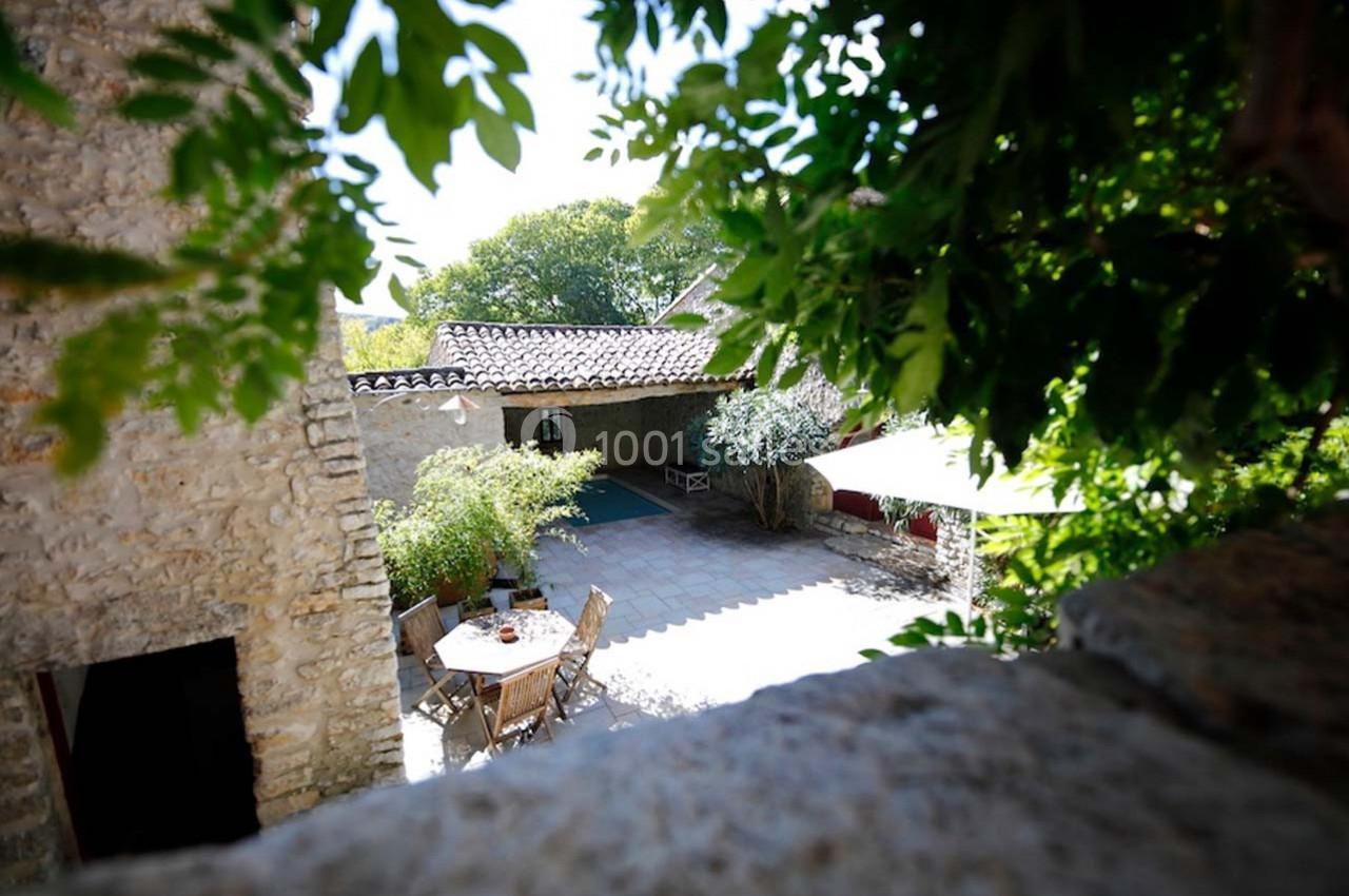 Cour ensoleillée avec table et chaises en bois, entourée de murs en pierre, végétation et arbres en arrière-plan.