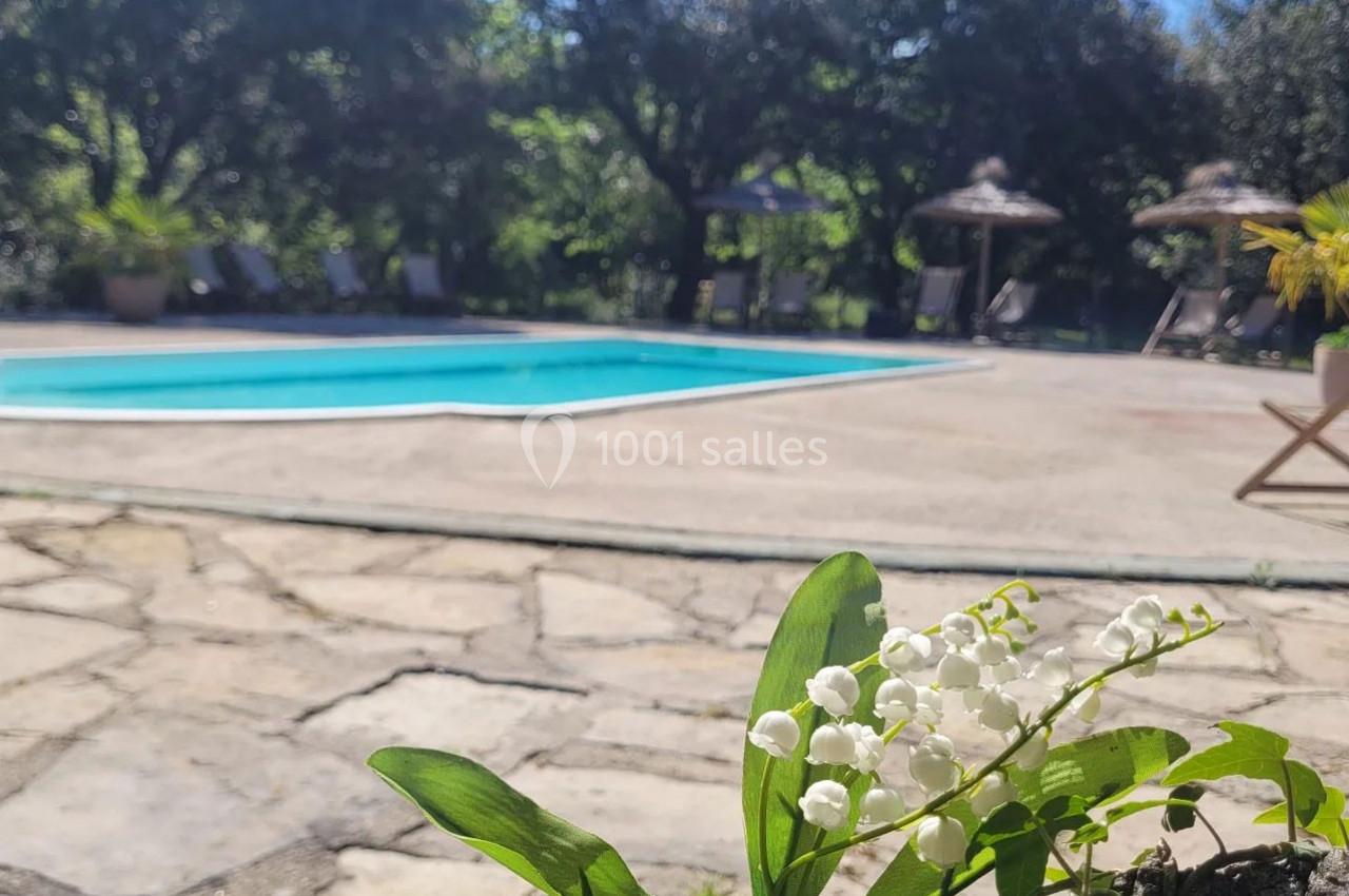 Muguet en premier plan devant une piscine entourée de chaises longues et de parasols dans un jardin arboré.