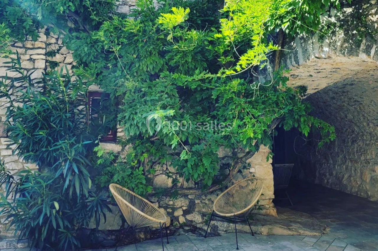 Mur en pierre recouvert de verdure avec deux fauteuils en osier disposés sur une terrasse pavée.