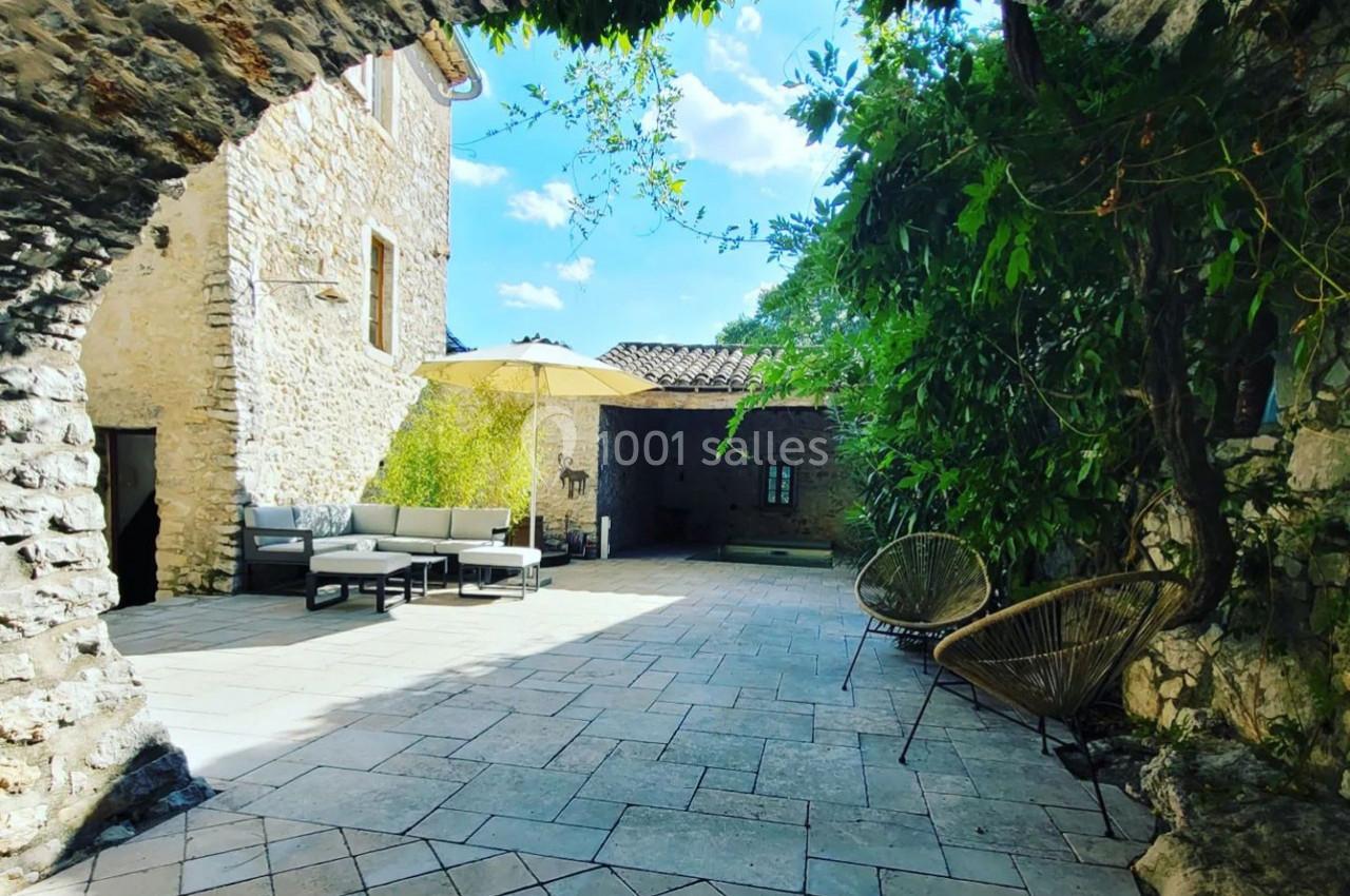 Cour en pierre avec mobilier de jardin, parasol et végétation, entourée de murs en pierre et vue sur le ciel dégagé.