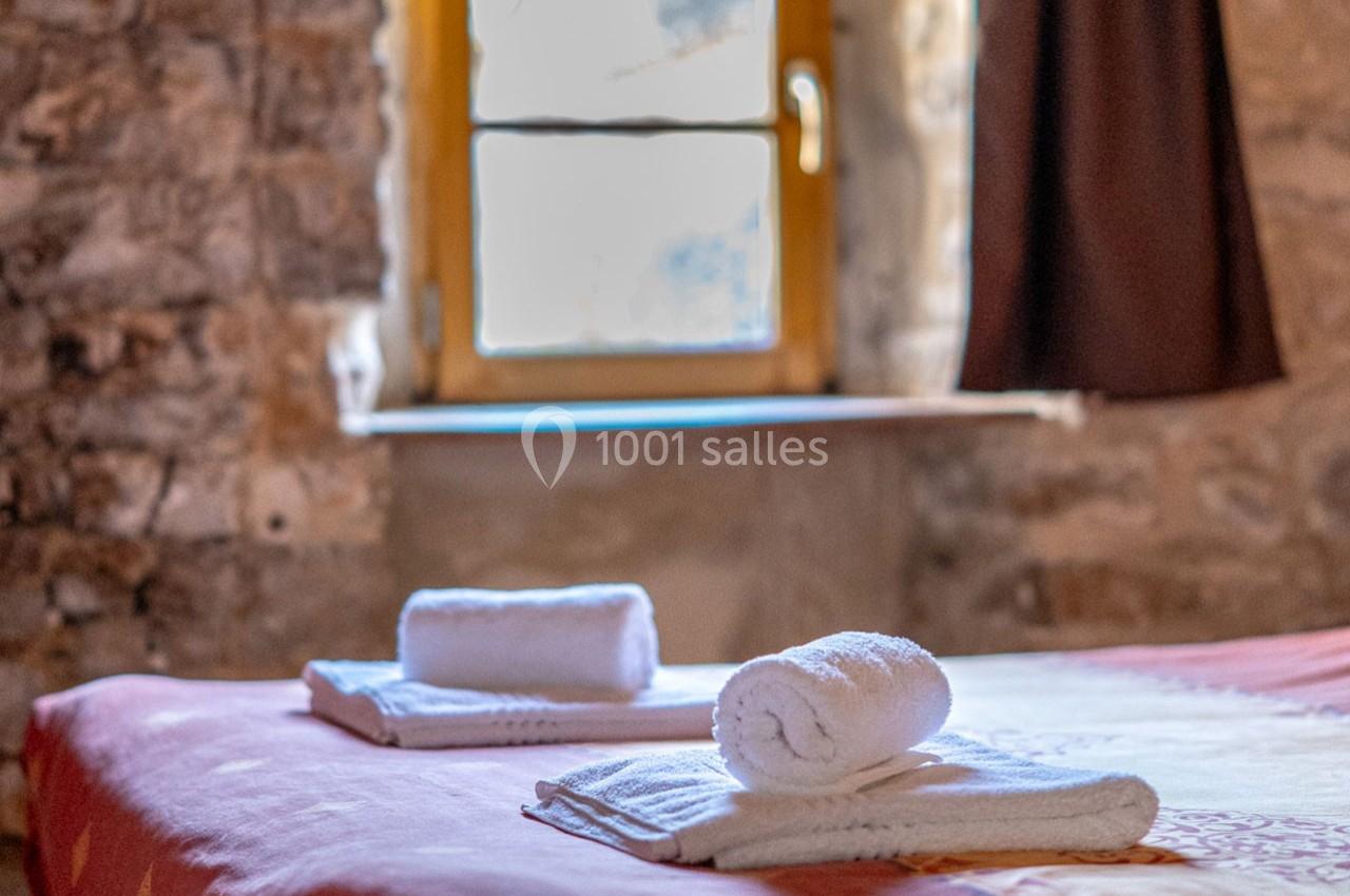 Deux serviettes blanches roulées posées sur un lit dans une chambre en pierre avec une fenêtre en arrière-plan.