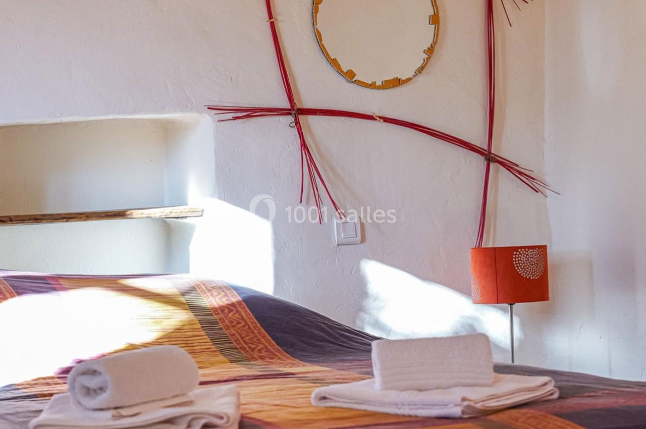 Lit avec couvre-lit coloré, serviettes pliées au premier plan, lampe orange et décoration murale en arrière-plan.