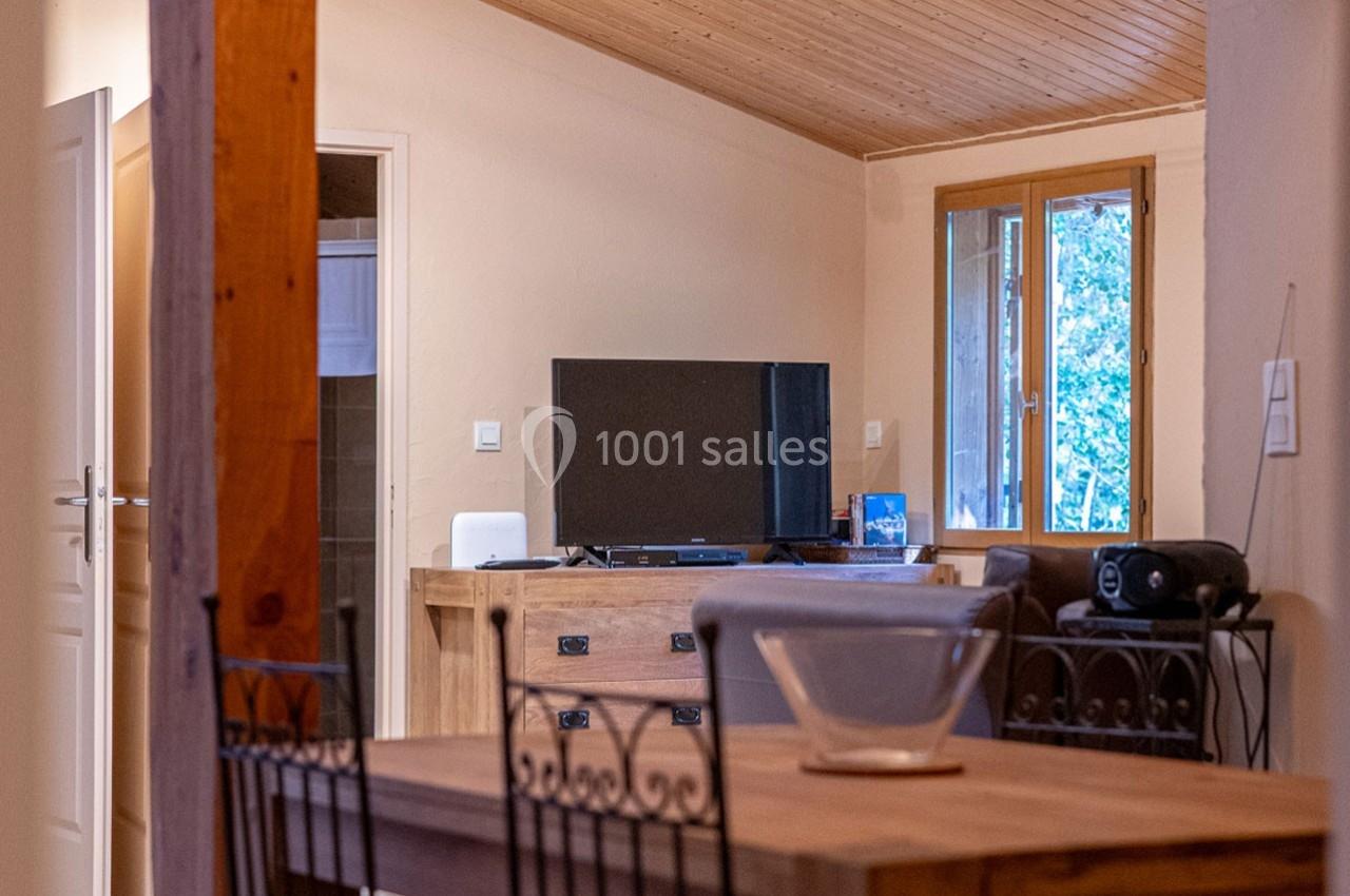 Intérieur d'une pièce avec table en bois, télévision sur un meuble, fenêtre donnant sur un extérieur arboré.