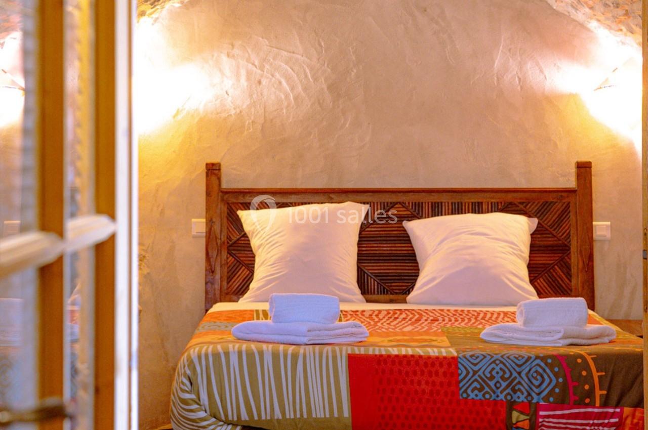 Lit double avec tête de lit en bois sculpté, draps colorés et serviettes pliées, dans une chambre aux murs en pierre.